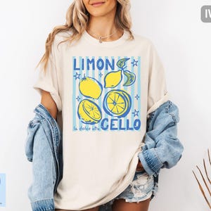 May include: Ivory-colored t-shirt with a graphic design featuring lemons, blue stripes, and the words "Limon Cello" and "la dolce vita." The design includes yellow lemons and blue stars. The shirt is a casual, oversized style.