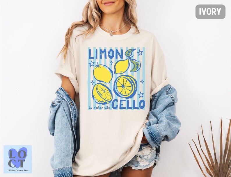 May include: Ivory-colored t-shirt with a graphic design featuring lemons, blue stripes, and the words "Limon Cello" and "la dolce vita." The design includes yellow lemons and blue stars. The shirt is a casual, oversized style.