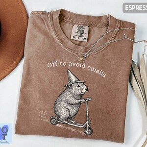 Puede incluir: Camiseta color espresso con el texto "Off to avoid emails" y un gr&aacute;fico de un roedor montado en un scooter y con un sombrero de mago. La camiseta tiene una etiqueta Comfort Colors.