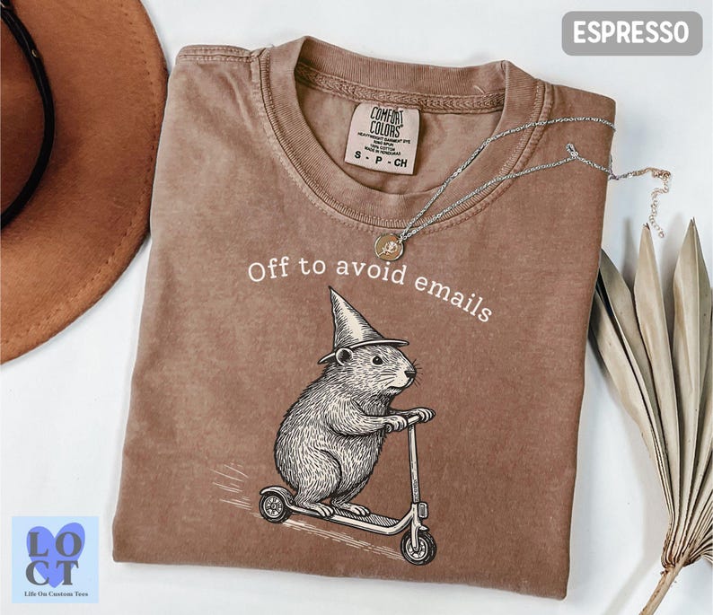 Puede incluir: Camiseta color espresso con el texto "Off to avoid emails" y un gr&aacute;fico de un roedor montado en un scooter y con un sombrero de mago. La camiseta tiene una etiqueta Comfort Colors.