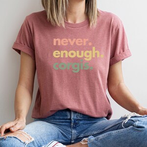 May include: A woman wearing a maroon t-shirt with the text "never. enough. corgis." printed in a rainbow gradient. She is sitting with her legs crossed and wearing blue jeans.