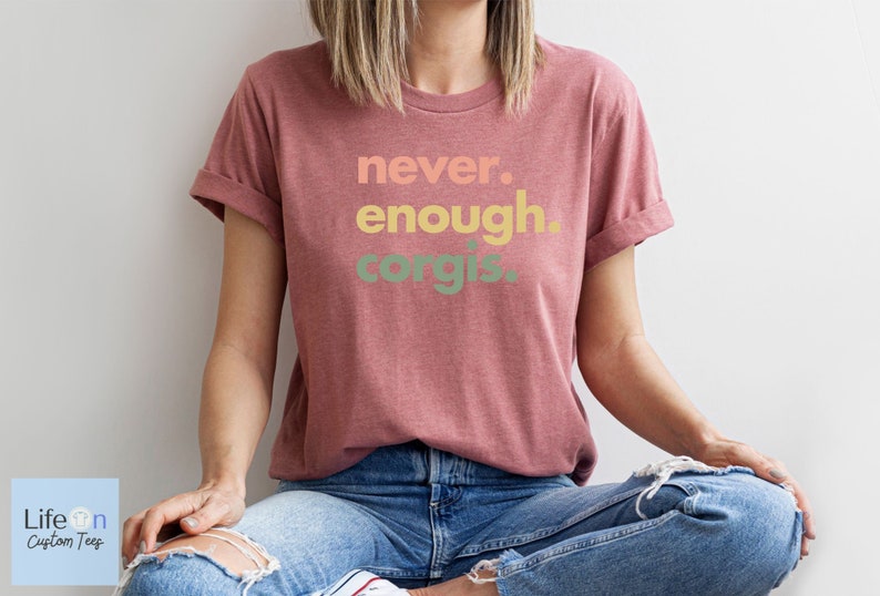 May include: A woman wearing a maroon t-shirt with the text "never. enough. corgis." printed in a rainbow gradient. She is sitting with her legs crossed and wearing blue jeans.