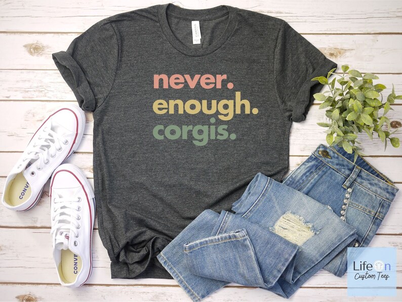 May include: A dark gray t-shirt with the text "never. enough. corgis." printed in colorful letters. The shirt is laid out on a white wooden surface with a pair of white sneakers and blue jeans.
