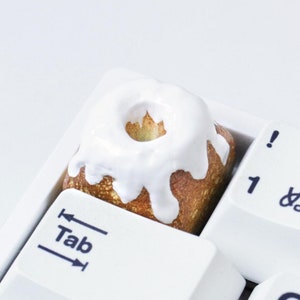 以下が含まれることがあります： 「Tab」キーに小さなケーキ型のキーキャップが付いた白いキーボード。ケーキは白いアイシングで覆われており、中心は茶色です。