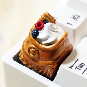 May include: A brown, fish-shaped taiyaki pastry keycap featuring a dollop of white whipped cream, a single red raspberry, and two small blueberries on top, installed on a white mechanical keyboard next to the Tab key.