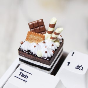 以下が含まれることがあります： 白いフロスティング、チョコレートスプリンクル、チョコレートバー、ストライプのワッフルスティック2本をトッピングしたミニチョコレートケーキ。ケーキは「Tab」キーが見える白いキーボードの上に置かれています。
