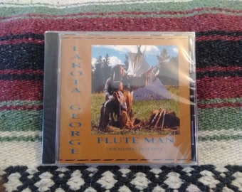 以下が含まれることがあります： Lakota Georgeのアルバム「Flute Man」のCDケース。カバーアートは、ネイティブアメリカンの衣装を着た2人の人物と、背景にティピーとキャンプファイヤーを描いています。アルバムタイトルとアーティスト名は青色のテキストです。