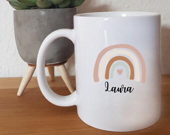 May include: White ceramic mug with a rainbow design in pink, beige, brown, and green. The rainbow has a small pink heart in the center. The name "Laura" is written in black below the rainbow.