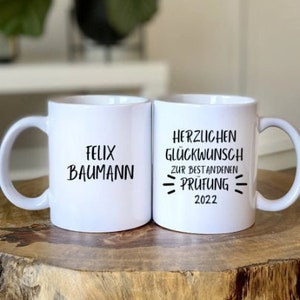 May include: Two white ceramic mugs with black text. The mug on the left says "Felix Baumann". The mug on the right says "Herzlichen Glückwunsch zur bestandenen Prüfung 2022".