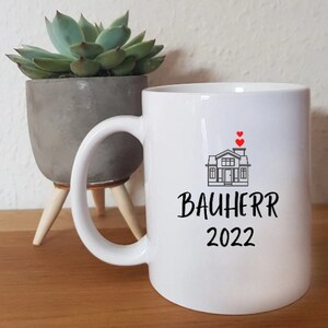 Puede incluir: Taza de cerámica blanca con una ilustración en blanco y negro de una casa con corazones encima. El texto "Bauherr 2022" está impreso en negro debajo de la casa.
