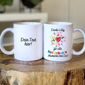 Puede incluir: Dos tazas de cerámica blanca. Una taza tiene el texto "Dein Text hier!" y la otra taza tiene el texto "Danke Lilly, für alle kunterbunte Momente mit Dir!" con un diseño de huellas de manos de colores.
