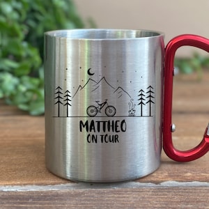 Könnte beinhalten: Eine Edelstahl-Reisebecher mit einem roten Karabinergriff. Der Becher hat ein schwarz-weißes Design mit einer Bergszene mit einem Fahrrad, einem Lagerfeuer und Sternen. Der Text "MATTHEO ON TOUR" ist auf den Becher gedruckt.