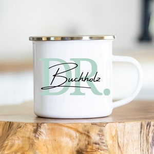 Peut inclure: Mug de camping en émail blanc avec un bord et une poignée argentés. Le mug présente un "DR" vert avec le texte "Buchholz" en écriture noire.