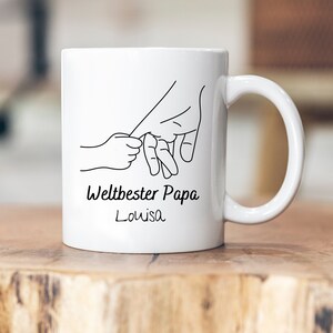 Op de afbeelding: Witte keramische mok met een zwarte lijntekening van twee handen die elkaars vingers vasthouden. De tekst "Weltbester Papa Louisa" is onder de tekening gedrukt.