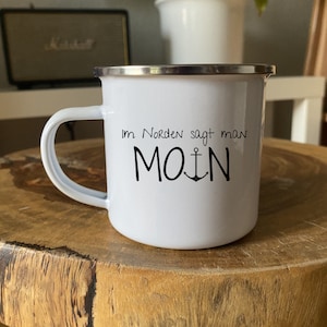 Puede incluir: Taza de esmalte blanco con borde plateado y diseño de ancla negra. La taza dice "Im Norden sagt man MOIN" en texto negro.
