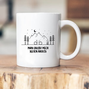 Könnte beinhalten: Weiße Keramiktasse mit einer schwarzen Bergszene, einem Halbmond, Sternen, einem Lagerfeuer und einem Fahrrad. Der Text "Man erlebt mich selten radlos" ist auf die Tasse gedruckt.