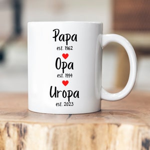 Peut inclure: Mug en céramique blanche avec du texte noir qui dit "Papa est. 1962", "Opa est. 1994", et "Uropa est. 2023". Chaque ligne de texte a un petit cœur rouge.