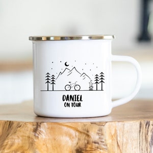 Op de afbeelding: Witte emaille campingmok met een zwart-witte illustratie van een berglandschap met een fiets, een kampvuur en sterren. De tekst "Daniel on Tour" staat onder de illustratie.