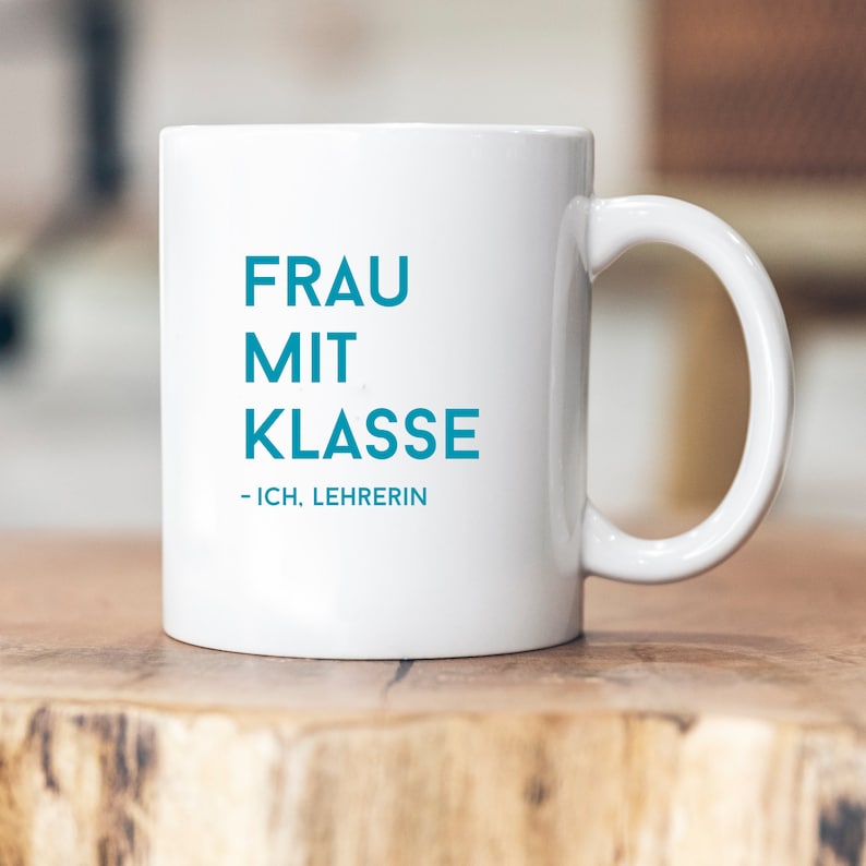 May include: White ceramic mug with a blue inscription that reads "FRAU MIT KLASSE - ICH, LEHRERIN".