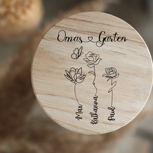 May include: A wooden round sign with the text "Omas Garten" and a heart symbol. Three stylized flowers with butterfly and names Max, Katharina, and Paul are engraved on the sign.