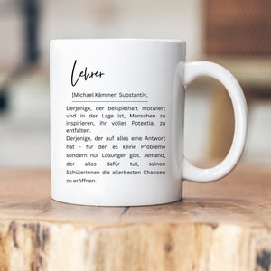 May include: White ceramic mug with a black and white design. The mug features the word "Lehrer" in a stylized font, followed by the definition of the word in German. The definition reads: "[Michael K&auml;mmer] Substantiv, Derjenige, der beispielhaft motiviert und in der Lage ist, Menschen zu inspirieren, ihr volles Potential zu entfalten. Derjenige, der auf alles eine Antwort hat - f&uuml;r den es keine Probleme sondern nur L&ouml;sungen gibt. Jemand, der alles daf&uuml;r tut, seinen Sch&uuml;lerinnen die allerbesten Chancen zu er&ouml;ffnen."