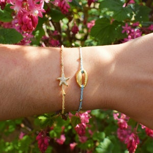 May include: Two bracelets on a wrist. One bracelet is light blue with a gold seashell charm. The other bracelet is beige with a gold starfish charm.