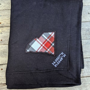 May include: Black throw blanket with a red and white plaid design of the state of Georgia and the coordinates "34.0007° N 81.0348° W" embroidered in white.