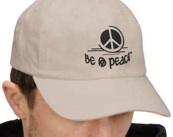 May include: A light beige baseball cap featuring a black graphic of a peace sign inside a circle above the text "BE D PEACE". The cap has a structured crown, a curved brim, and embroidered eyelets on the top panels for ventilation.