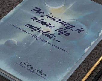 May include: A hardback journal with a blue, cloudy, and celestial-themed cover featuring the text "The journey is where life unfolds..." and "Sofia Rosa" in dark script, accompanied by two pencils and several gold-toned metal binder clips.
