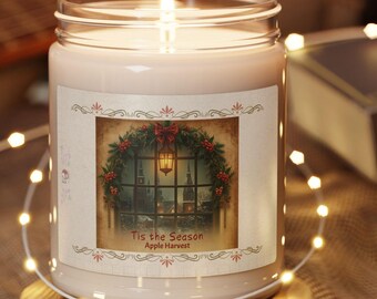May include: A clear glass jar candle with a lit wick, filled with off-white wax. The label features a festive window scene with a wreath, red berries, and the text 'Tis the Season Apple Harvest'. It sits on a round wooden base.