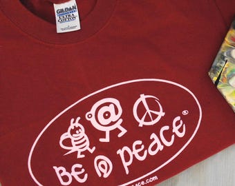 May include: A maroon t-shirt with a white oval design featuring a bee, a person, and a peace symbol. The words "be @ peace" are printed in white, along with the website be-at-peace.com. The shirt has a crew neck and short sleeves.