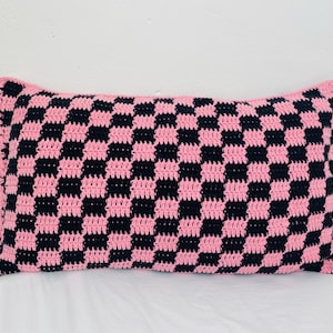 Op de afbeelding: Een roze en zwart geruit gehaakt kussen. Het kussen is gemaakt met een eenvoudig ruitpatroon.