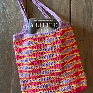 Op de afbeelding: Een gehaakte draagtas met paarse, roze en gele strepen. De tas bevat een boek getiteld "A Little Life" met de tekst "Man Booker Prize Finalist" op de kaft.