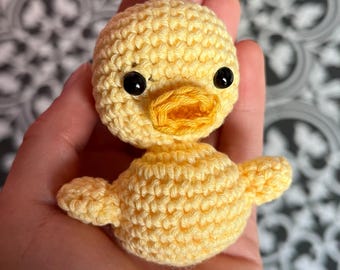 May include: A small yellow crocheted duck with black eyes and an orange beak.