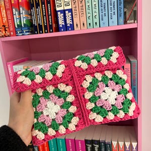 May include: A handmade, pink crochet clutch with a granny square design. The clutch features squares of white, green, and pink yarn. The bag is folded over and held open, revealing the interior. The background includes a pink bookshelf filled with books.