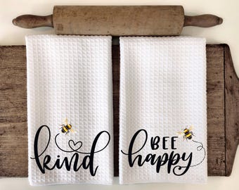 May include: Two white waffle weave kitchen towels with black text and bee illustrations. The first towel says "bee kind" with a heart and a bee. The second towel says "bee happy" with a bee.