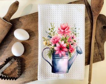 May include: A white kitchen towel with a watercolor design of pink flowers in a blue watering can. The flowers are surrounded by green leaves and stems.