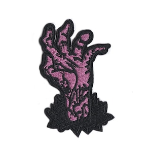 Op de afbeelding: Geborduurde patch met een paarse zombiehand die uit een zwarte, gekartelde basis komt. De skeletdetails van de hand zijn fijn geborduurd. Ideaal voor jassen, tassen of andere textielwaren.