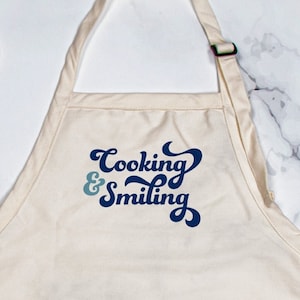 May include: Cream-colored apron with the text "Cooking & Smiling" in blue script. The apron has three front pockets and an adjustable neck strap. Wooden spoon and slotted spoon are on the sides.