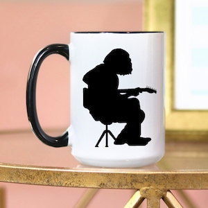 Può includere: Tazza in ceramica bianca con manico e bordo neri. La tazza presenta una silhouette nera di una persona che suona la chitarra seduta su uno sgabello. La tazza è su una superficie dorata.