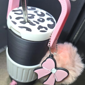 May include: A black and white tumbler with a pink handle and a pink bow charm. The tumbler has a black and white leopard print lid. A fluffy pink pom-pom is attached to the charm.