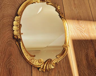 May include: An oval wall mirror featuring an ornate gold-colored frame with intricate scrollwork and leaf-like carvings along the edges. The reflective glass surface is smooth and clear, capturing light and surrounding details within its oval shape.