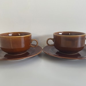 May include: Two brown ceramic soup bowls with matching saucers. The bowls have a rounded shape and a slightly flared rim. The saucers are plain and have a slightly raised edge.