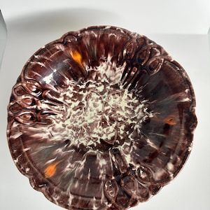 Op de afbeelding: Een ronde keramische kom met een glanzend glazuur in een gemarmerd patroon van donkerbruin, crème en wit. De rand heeft een geschulpte vorm met reliëf ovale motieven. Het midden vertoont een textuurrijke, gevlekte afwerking met oranje accenten.