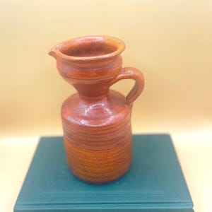 Può includere: Un piccolo vaso in ceramica arancione con un'ampia apertura e un manico. Il vaso ha una superficie testurizzata ed è appoggiato su un libro verde.