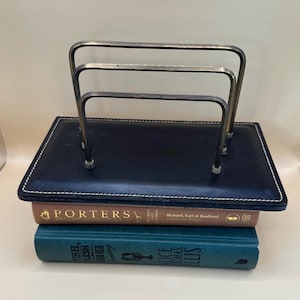 Può includere: Un organizer da scrivania in pelle blu navy con un portalettere in metallo argentato. L'organizer è rettangolare con cuciture bianche. Due libri sono impilati sotto.