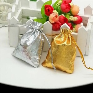 Peut inclure: Deux petits sacs cadeaux à cordon. L'un est argenté, l'autre doré. Les deux sacs sont faits d'un matériau brillant et texturé. Les sacs sont noués en haut avec des cordons assortis. Le fond comprend une barrière blanche et des fleurs.