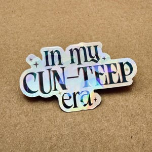 Puede incluir: Una pegatina holográfica con forma troquelada que presenta el texto negro "in my CUN-TEEP era" en una fuente serif negrita. La superficie refleja un espectro de colores e incluye pequeños iconos de estrellas dispersos alrededor de las letras.