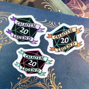 May include: Three die-cut stickers featuring a black, geometric D20 design with the text "CHAOTIC 20 LEGEND" on a banner. Each sticker has a different color scheme: purple, orange, and pink. The stickers are on a dark blue surface.