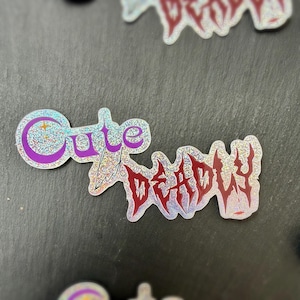 以下が含まれることがあります： 「Cute but Deadly」の文字が入ったホログラフィックステッカー。「Cute」の文字は、キラキラした背景に紫色のバブルフォントで書かれています。「Deadly」の文字は、赤いギザギザのフォントで書かれています。